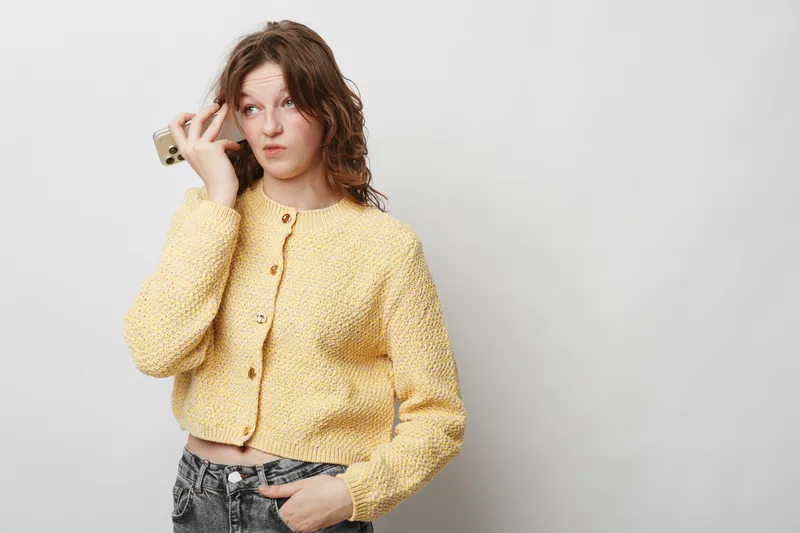 Young woman in yellow knitted sweater holds smartphone near ear, looking confused and thoughtful. Isolated on white background. Concept of communication, doubt, and decision making.