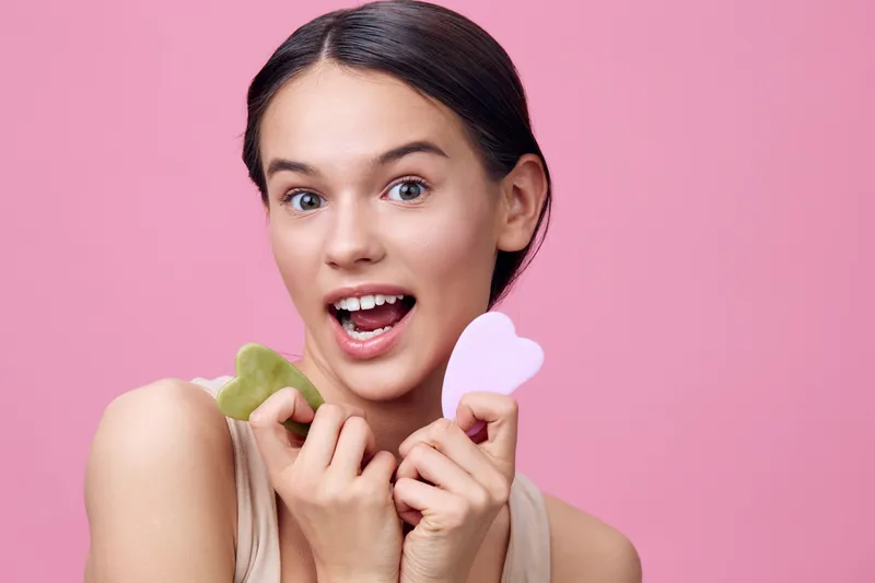 Young woman holding facial tools, showcasing skincare routine, smiling against a pink background The image emphasizes beauty and self care concepts