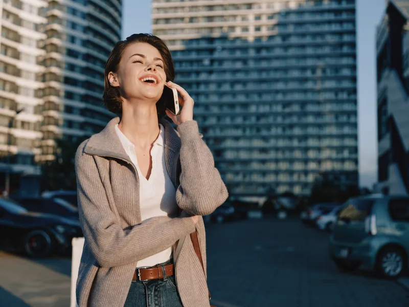 A cheerful professional woman is seen talking on a cellphone in an urban outdoor setting. She smiles and looks engaged, creating a positive and dynamic atmosphere.
