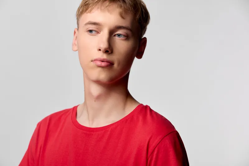 Young man in a red shirt gazes to the side with a thoughtful expression, studio lighting and a minimal background create a clean lifestyle image for fashion, branding, or editorial use