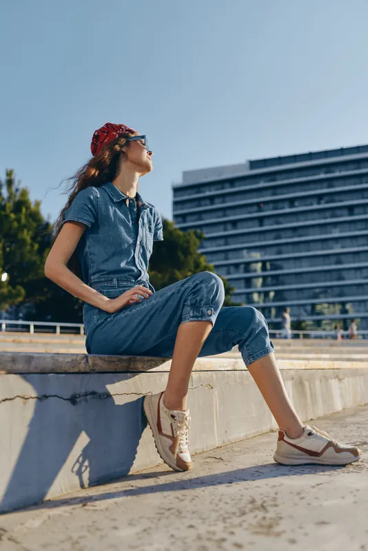 young woman wearing jumpsuit and sneakers relaxing outdoors in urban setting under sunlight during daytime