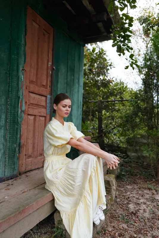 Calm portrait of a woman in a pale yellow dress sitting on a doorstep outside a wooden green door, surrounded by trees, creating a serene and contemplative outdoor fashion mood with natural light