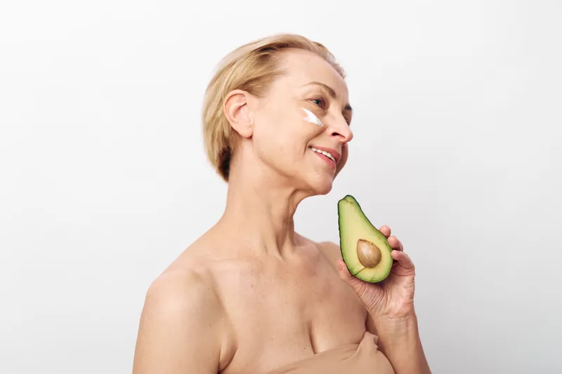 Mature woman with short hair applies natural cream under eyes and holds fresh avocado half. Skincare concept for healthy aging, natural beauty, and organic facial care on white background.