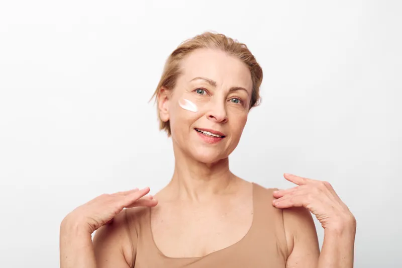 Mature woman with light skin applies face cream on cheek, smiling gently. Soft natural makeup and beige top against white background. Concept for skincare and aging beauty care.