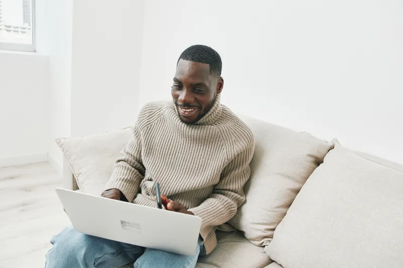 Smiling Black man working on laptop in cozy sweater, sitting on couch at home, creating a productive atmosphere and embracing remote work lifestyle