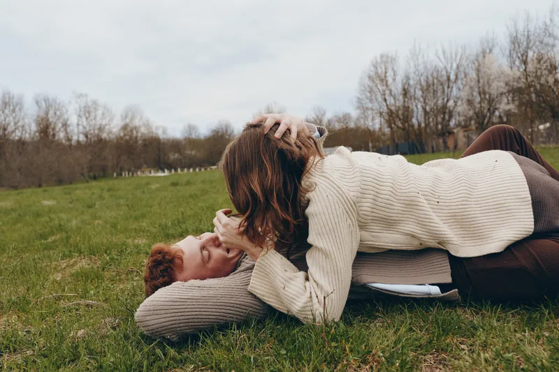 couple affection outdoors friendship laughter on the grass, a woman and a man share a playful moment in cozy sweaters, under a clear sky, showcasing casual lifestyle, outdoor leisure, and genuine