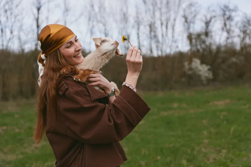 A joyful woman in a brown coat and orange headscarf lifts a smiling goat kid outdoors, offering a small flower. A candid lifestyle moment celebrating bond with animals in nature.