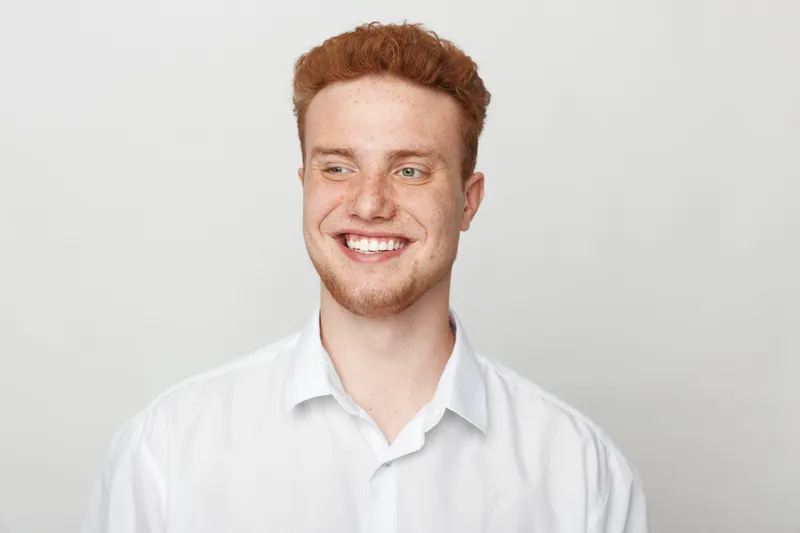 Smiling young man with red hair and freckles wears white shirt, looking away with happy expression. Studio portrait on light background. Concept of youth, confidence, and positive mood.