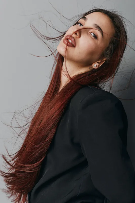Woman with long red hair and dark lipstick posing in black jacket against gray background. Fashion portrait with wind effect and confident expression on female face.