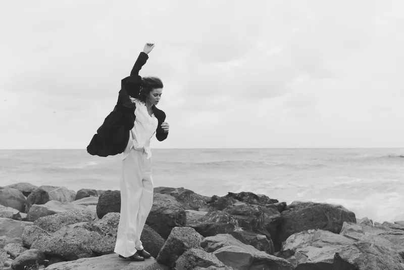 Woman on a rocky coast in wind, raising her arm in motion against the ocean and sky, outdoor fashion scene, monochrome image of strength, balance and resilience with scarf and coat, dramatic seascape