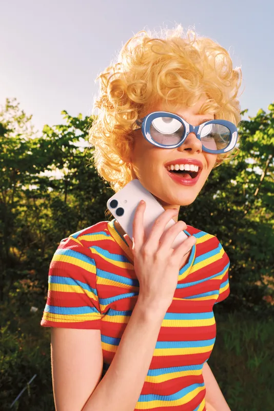 A joyful woman with retro style, blonde curls and oversized glasses chats on a phone outside in bright sunlight, capturing casual lifestyle and playful vintage fashion.