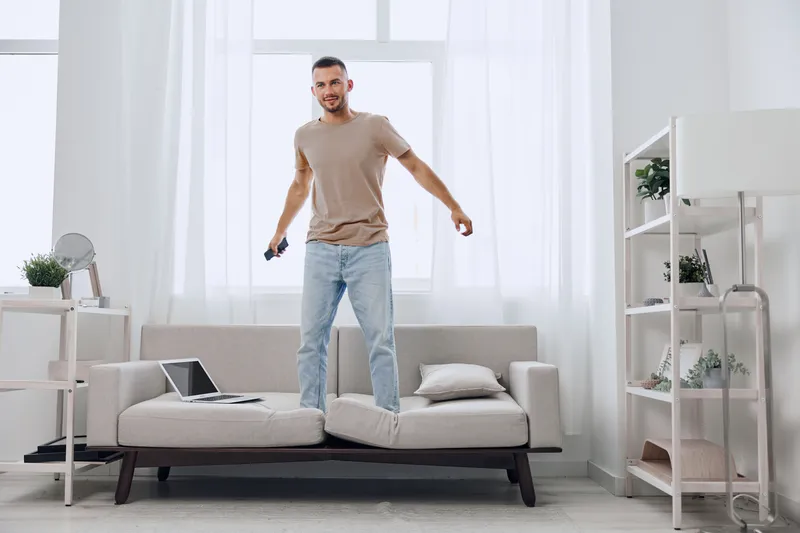 Energetic man joyfully jumping on a sofa, showcasing vibrant lifestyle with a laptop nearby. Bright, modern interior creates a lively atmosphere for home activities.