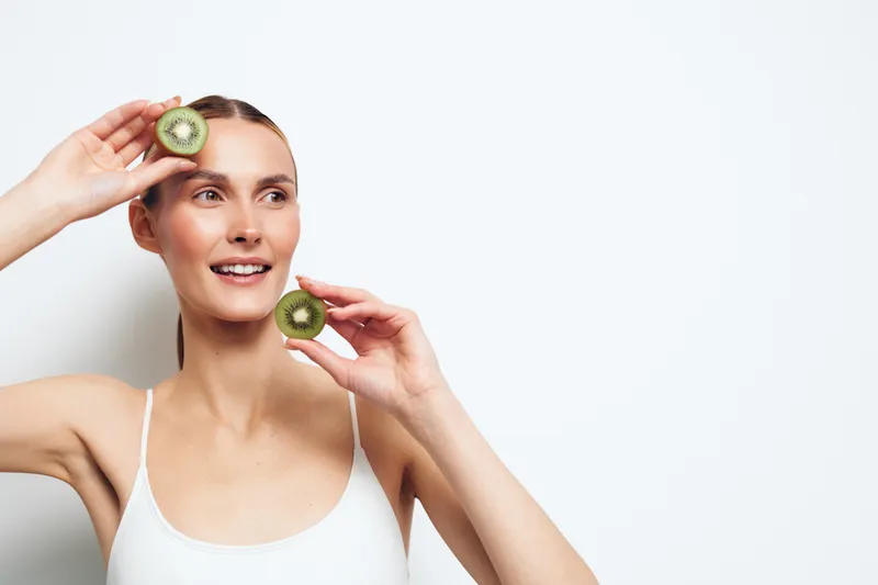 Woman holding kiwi slices for skincare on face beauty spa self care freshness healthy wellness natural mask treatment smiling in clean white studio soft light