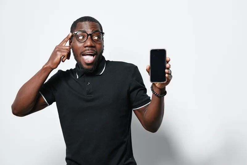 African American man with dark skin and glasses showing smartphone with blank screen, pointing to head with surprised expression on white background in casual black shirt.