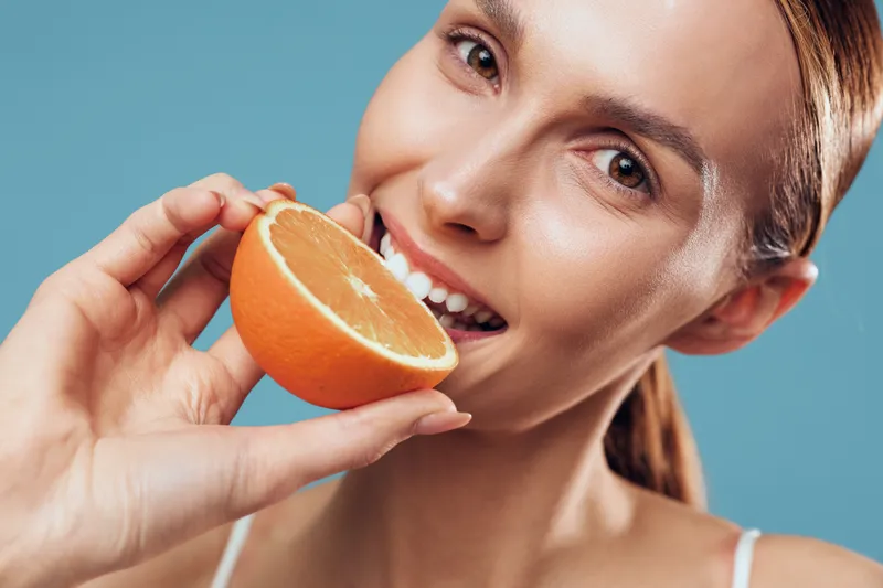 Closeup portrait of smiling woman holding half an orange near her mouth, healthy skin, natural light, fresh fruit, blue background, positive mood, wellness concept