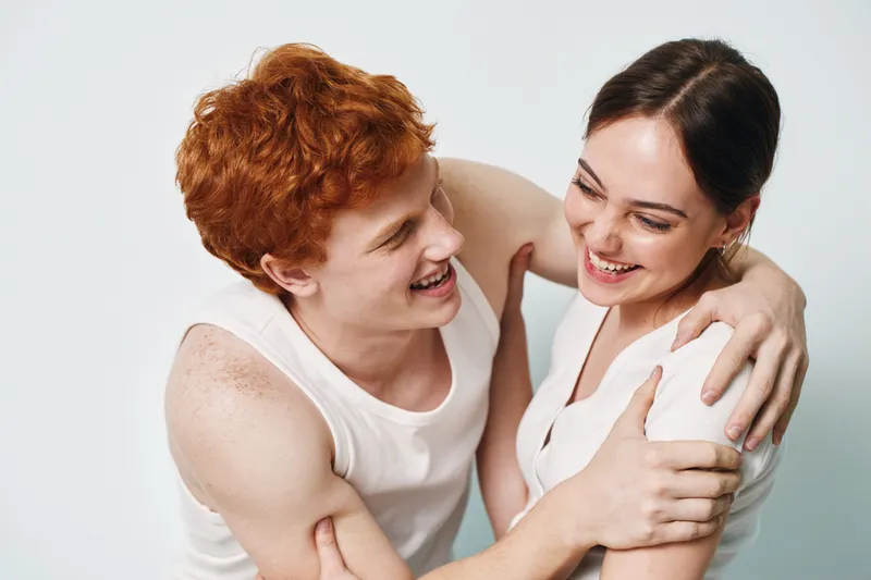 Young man and woman laughing together in casual white tank tops with friendly interaction and happiness on light isolated background. Closeup, joyful people enjoying moment with smiles and embrace.