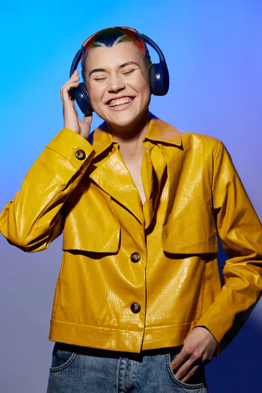 Smiling woman in yellow jacket enjoying music with headphones, vibrant background, casual style, joyful expression, contemporary urban vibe.