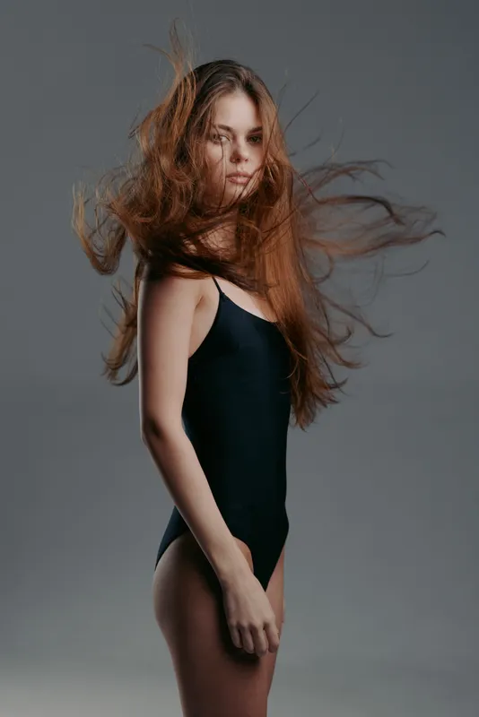 Young woman with flowing hair wearing black bodysuit posing in studio with gray background, elegant and dynamic beauty concept