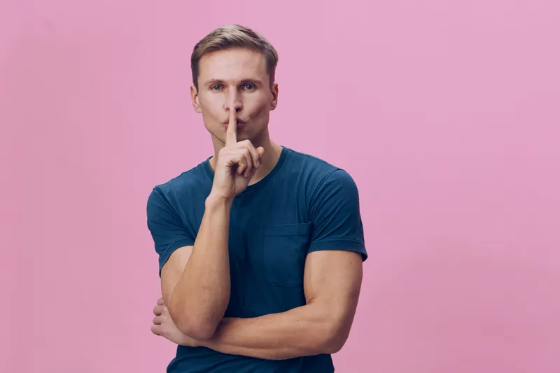 Confident man with blond hair in casual blue t-shirt holding finger to lips gesture isolated on pink background studio portrait. People lifestyle concept, quiet, secret, silence, discreet.