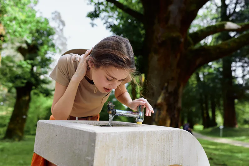 Girl child drinking water from public fountain in park, leaning over concrete basin in casual summer clothing, outdoor scene with trees and greenery, hydration and playful moment.