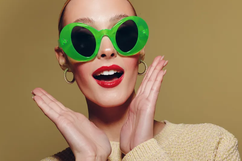 Young woman with bright red lipstick and large green sunglasses poses with hands near face on beige background. Concept for fashion, beauty, and summer style portrait.