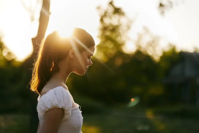 A young woman with long hair in a white sundress standing in the golden hour light, showcasing a serene expression and nature backdrop in soft warm tones
