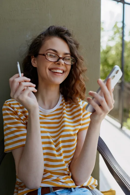 A smiling woman in a yellow and white striped t shirt sits on a balcony holding a phone and cigarette, enjoying a sunny moment with relaxed casual style.