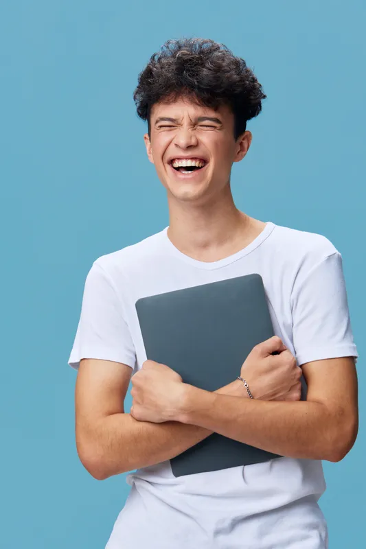 Laughing young man holding laptop, wearing white t-shirt with curly hair, standing against plain blue background, expressing joy and happiness.