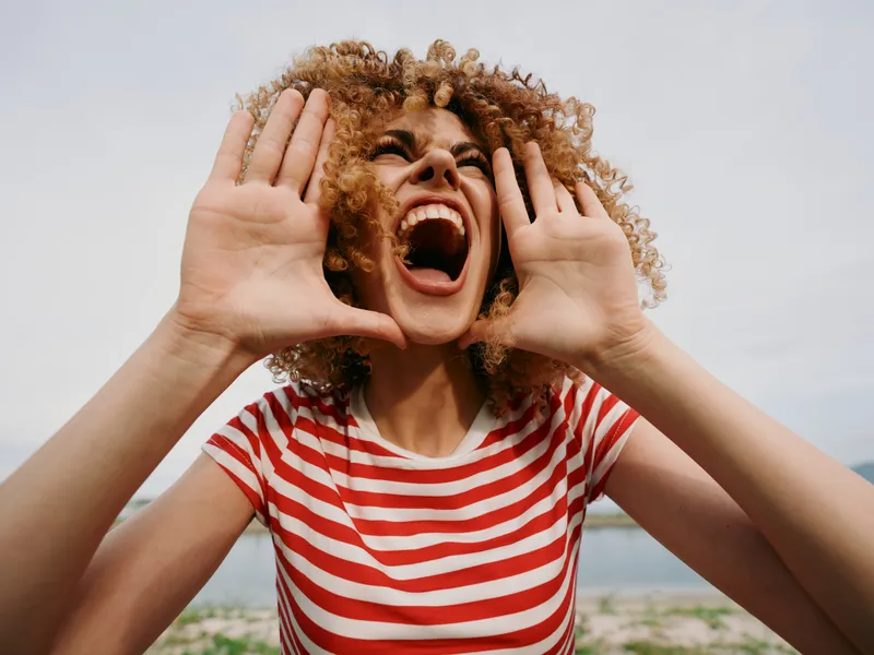 Vibrant portrait of a young woman at the beach, shouting with hands near her face, wearing a red and white striped shirt under a bright sky and ocean breeze.