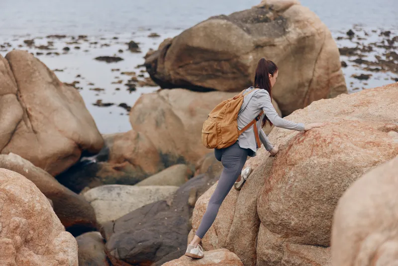 Young woman climbing rocky terrain near the sea, wearing athletic gear and a backpack, showcasing adventure and outdoor spirit