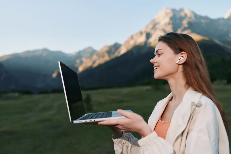 A cheerful professional works outdoors with a laptop, smiling as she types, standing in a bright field with distant mountains. The scene conveys optimism, focus, and tech enabled productivity in