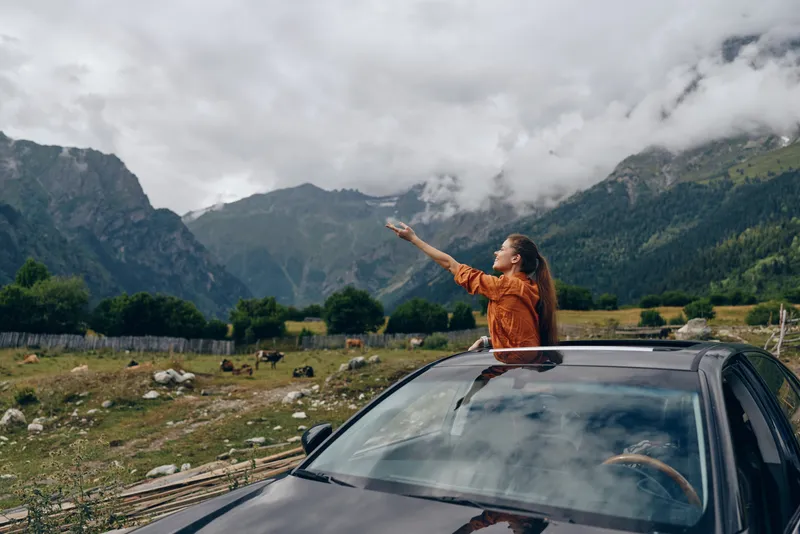 A traveler in an orange jacket extends an arm from a car sunroof, driving through a dramatic mountain valley, capturing a wild sense of travel