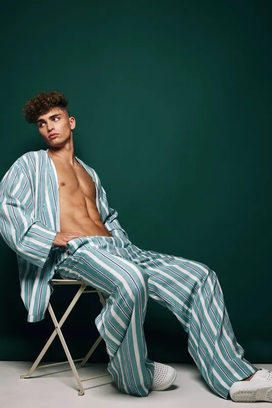 Young man with curly hair wearing striped loose shirt and pants sitting on white chair against green background. Casual fashion, relaxed pose and modern style with white sneakers.