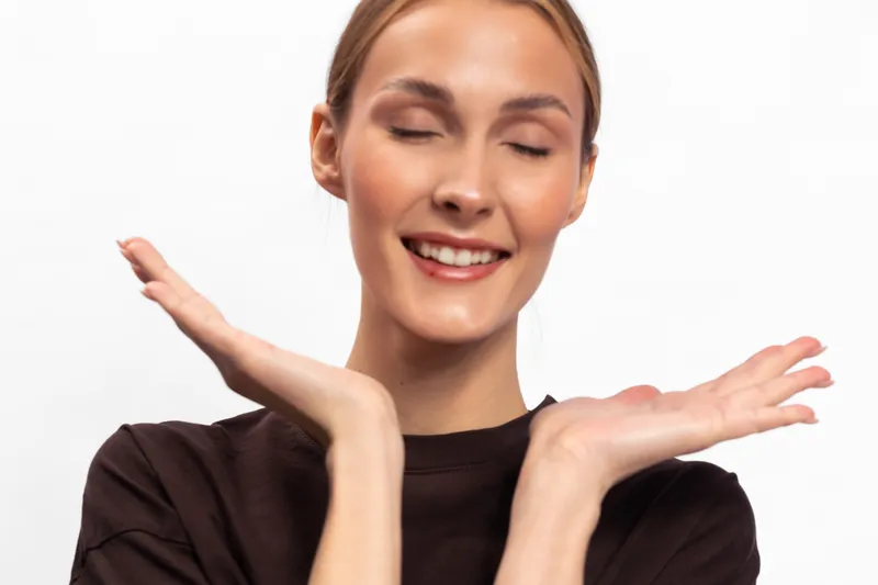 Smiling young woman with closed eyes and relaxed expression holds hands near face on white background. Concept of happiness, peace, and positive emotions with natural beauty and joy.