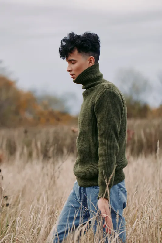 A young man in an olive turtleneck sweater walks slowly through tall dry grass under a muted overcast sky, a candid portrait captured in soft natural light emphasizing texture, authenticity and quiet