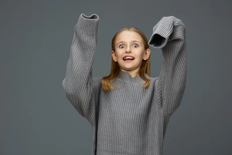 A joyful girl in an oversized gray sweater with excited expression, standing against a neutral gray background, perfect for portraying happiness and comfort