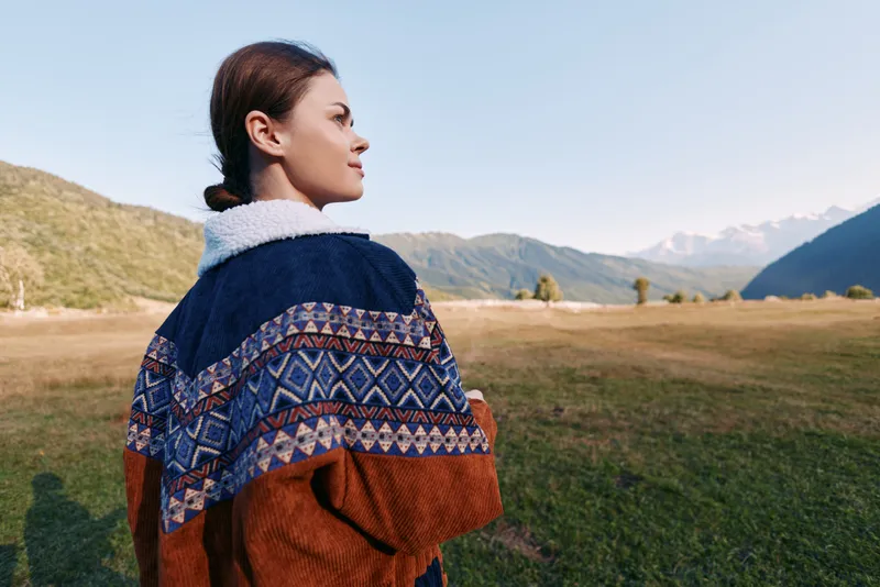 Woman in patterned jacket stands in a mountain field outdoors, portrait side view of young female looking toward horizon over green meadow, serene landscape with warm daylight and calm mood.