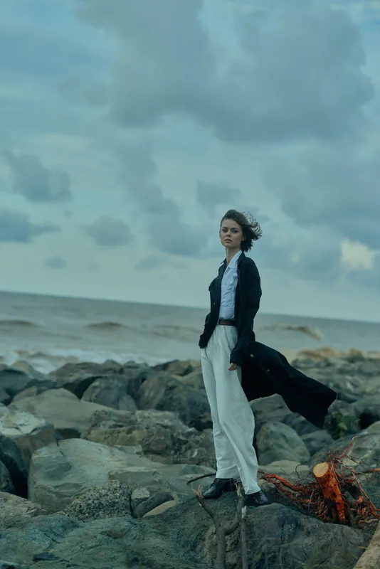Woman stands on rugged rocks by the sea, wind in her coat, cloudy sky above, creating a dramatic outdoor fashion moment along the coast