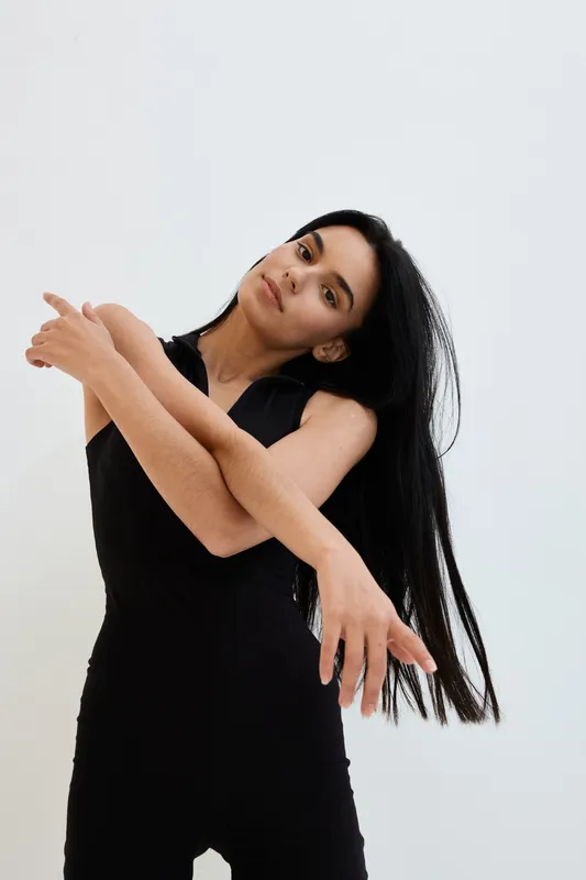 Young woman with long black hair posing gracefully in black sleeveless outfit against white background, expressing calm and elegance in minimalist style photography.