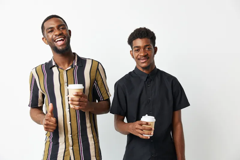 Two smiling African American men enjoying coffee, showcasing friendship and happiness in a modern setting. Their casual outfits radiate positive energy and warmth.