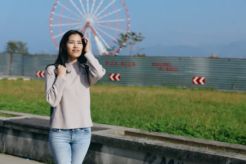 Woman stands outdoors near a ferris wheel wearing a light sweater and jeans smiling softly with hand in hair in a park setting on a sunny day for a casual lifestyle shot