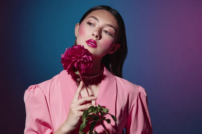 Fashion model with wet dark hair holds a bright magenta flower close to her face. She wears pink clothing and matching artistic makeup under soft colorful studio lighting. Beauty concept.