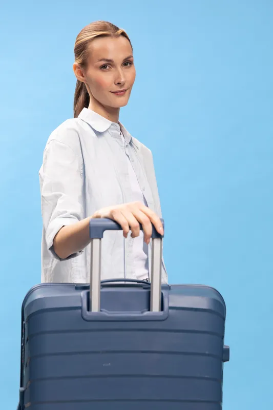 Young woman with light brown hair holds blue suitcase handle standing against blue background. Casual travel concept with confident facial expression and neat hairstyle in bright studio lighting.