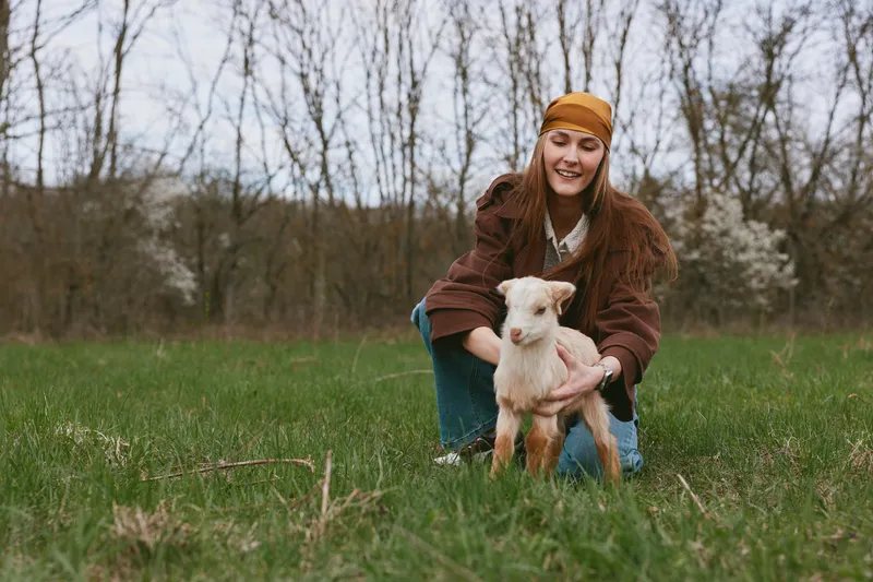 Smiling woman with a young goat outdoors, kneeling in a field, embracing the animal. Concept of rural lifestyle and gentle care. Natural daylight creates a warm mood in a countryside setting.