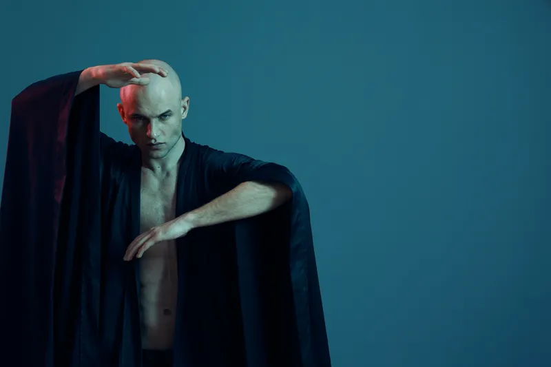 Captivating man in a flowing black robe striking a dramatic pose against a monochromatic blue background, showcasing elegance and creativity in a bold manner.