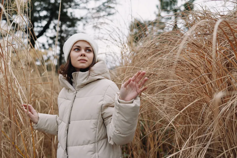 Woman in a light winter coat explores tall grasses outdoors, posing with a calm expression, serene natural backdrop, chilly air and soft light in a winter mood
