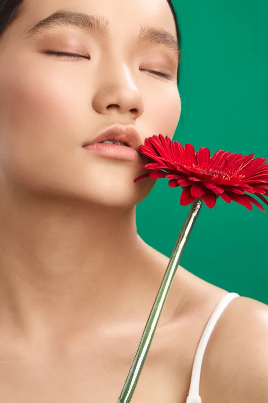asian woman rests with eyes closed, holding a red flower near her lips, a vibrant green background enhances a serene beauty portrait with soft light and gentle color contrast.