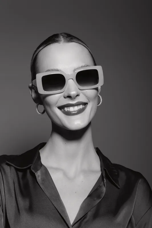 Stylish caucasian woman with pale skin smiles wearing oversized sunglasses and hoop earrings in studio. Fashion, beauty, glamour concept with confident mood.