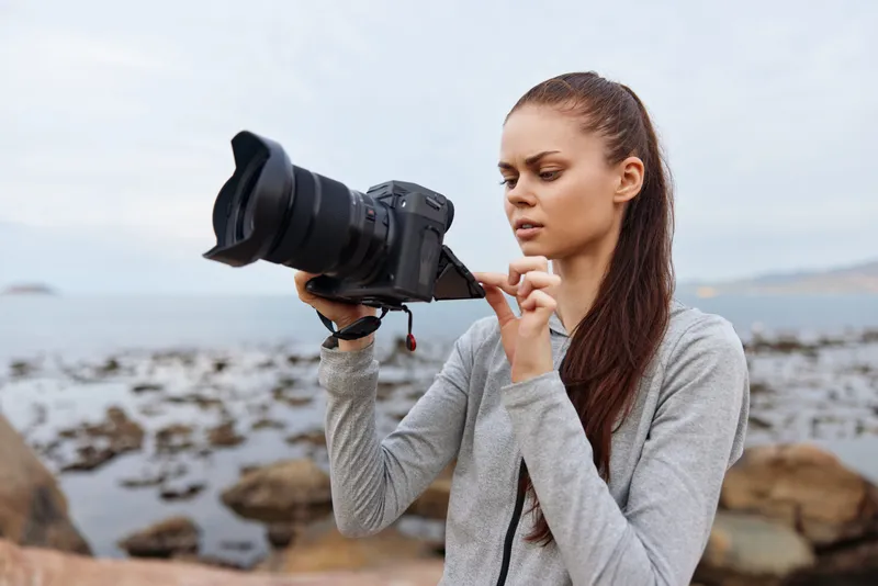 Young woman with long hair capturing scenery with camera on rocky beach, showcasing passion for photography and outdoor exploration