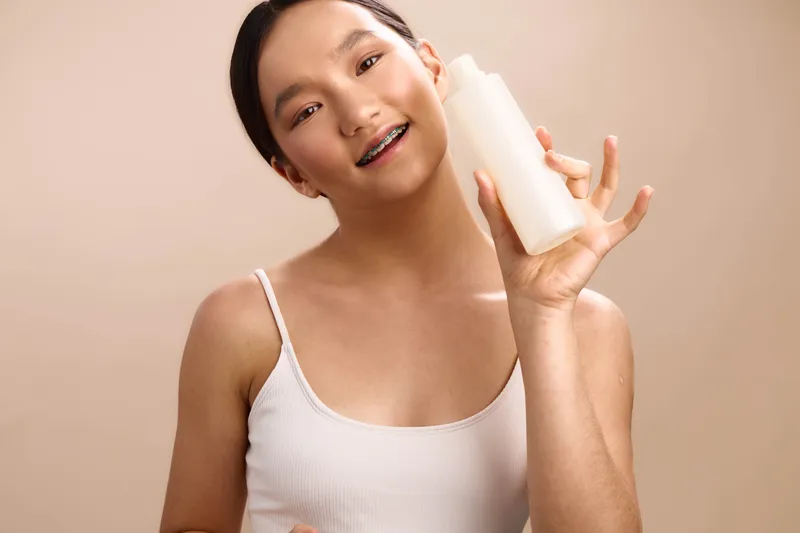Asian woman in a white tank top holds a skincare bottle and smiles at the camera, presenting a fresh, healthy beauty moment in a clean, modern studio setting.